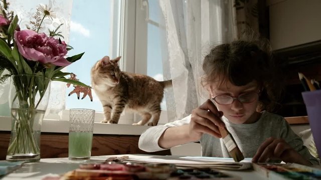adorable little girl painting at the album with a cat on background. young painter in creativity process in cozy sunny room