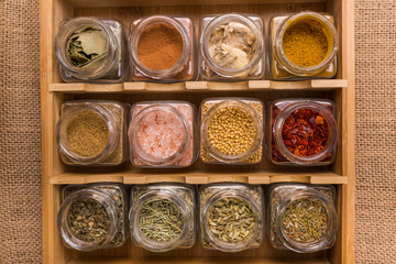 spices and aromatic herbs of different colors and textures in lined glass jars, on a brown background, with a wooden spoon
