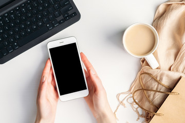 Female hands holding smartphone with blank screen mockup over home office desk with laptop, coffee cup, blanket. Shopping online at mobile phone, e-commerce concept