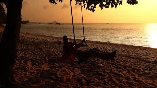 Man sitting and swinging on wooden swing at the seashore. Slow motion of young man teeting one way at the sandy shore of the ocean at the sunset. Theme of vacation and travelling in Thailand.