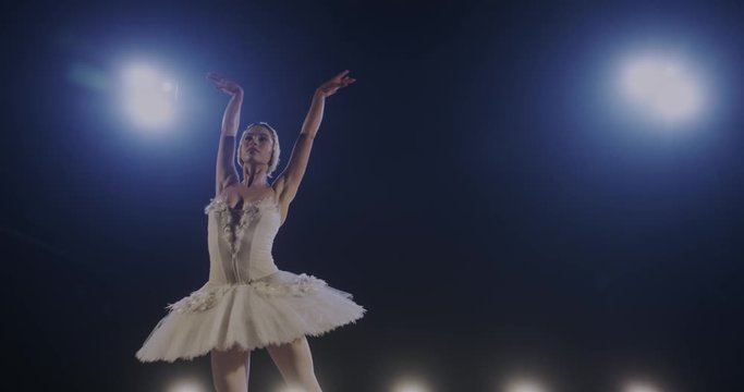 Slow motion of diligent young slim female ballet dancer in white dress dancing classical pas in blue light of lamps Beautiful ballerina hard-working and rehearsing perfomance of play alone in studio.