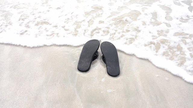 Black slippers on the beach touched by waves from the sea. slow motion.