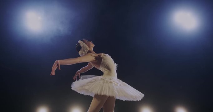 Pretty young ballerina leaning on side and doing dancing pas in blue spotlight on stage during performance. Classical ballet dancer in white tutu training and working hard. Perfect dance concept.