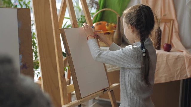 Teacher at art Studio teaches children, the girl sitting in front of his easel carefully listens to the teacher.