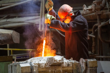 worker with hard hat and face mask stirs liquid metal in a furnace by steel bar