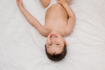 Cute toddler boy in diapers lies on bed with white sheet