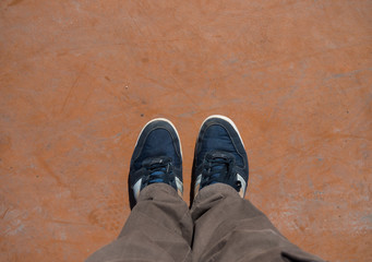 Top view of male legs in brown pants and blue sneakers on an orange concrete floor.