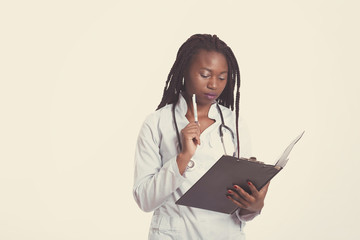 Fototapeta premium Female american african doctor, nurse woman wearing medical coat with stethoscope write test results in hospital sheet chart. Serious excited for success medical worker posing on light background
