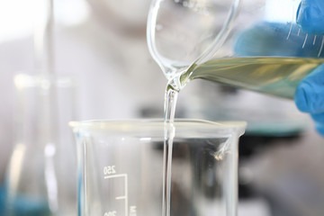 Scientist arm wearing blue protective gloves pouring yellow liquid at laboratory flask