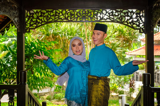 A portrait of young couple of malay muslim in traditional costume during Aidilfitri celebration showing welcome gesture by traditional wooden house