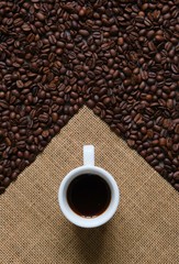 A white Cup of coffee on a background of coffee beans and coarse burlap. Coffee composition.