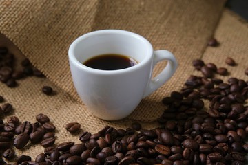 A white Cup of coffee on a background of coffee beans and coarse burlap. Coffee composition.