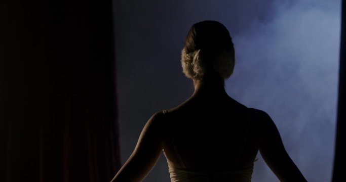 Theater curtains opens and Caucasian graceful ballerina in tutu entering the stage and doing bowing pas for audience. Back view on female ballet dancer coming out and bow in darkness. Rear.