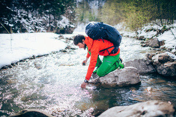 Young male tourist fillings water into the bottle from a river in the mountains..Beautiful winter landscape with snow covered banks and trees  on background. Climbing, trekking, active life concept.
