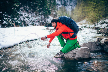 Young male tourist fillings water into the bottle from a river in the mountains..Beautiful winter landscape with snow covered banks and trees  on background. Climbing, trekking, active life concept.