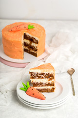 Birthday cake and piece of cream cake with carrot paste, stack of white saucers, cake stand, pink background, white table, vertical format.