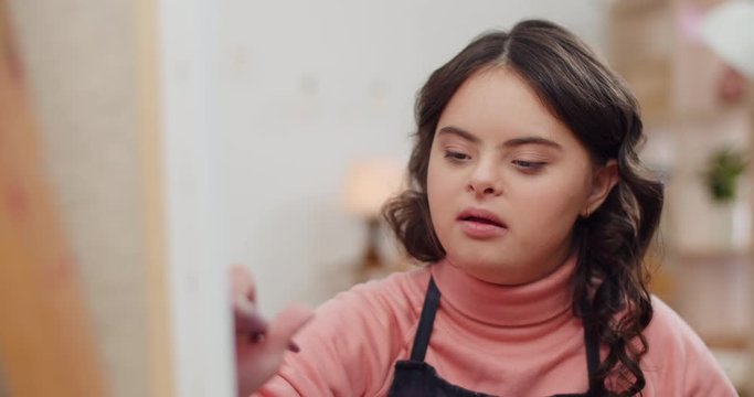 Close up view of talanted girl with down syndrome in painting picture and smiling.Beautiful teenager looking happy while sitting in front of molbert . Concept of sunny children. Indoors.