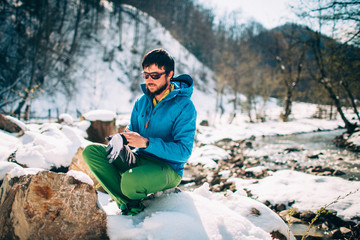 Young male tourist puts on gloves near a river in the mountains..Beautiful winter landscape with...