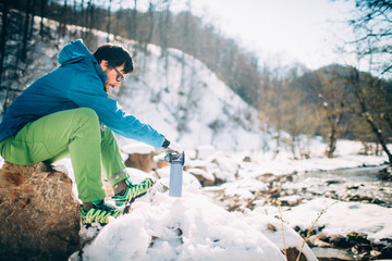 Fototapeta na wymiar Young male tourist drinks hot coffee from a thermos near a river in the mountains. Beautiful winter landscape with snow covered banks and trees on background. Climbing, trekking, active life concept