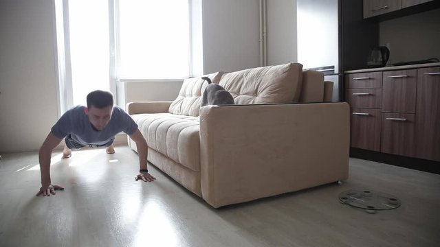 Strong Fitness Men in Athletic Workout Clothes. He is Training at Home in Her Living Room. The cat sits on the couch and watches the owner