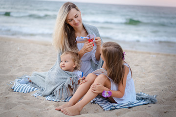 Fototapeta na wymiar Mom with daughters blondes in white dresses laugh, hug, draw and cut out of paper and sit near the blue sea on the beach at sunset and hide behind a gray knitted blanket.