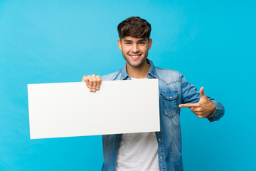 Young handsome man over isolated blue background holding an empty white placard for insert a concept