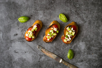 Three italian bruschetta with avocado, feta cheese and tomatoes. Basil leaves on the grey background and a steel knife. 