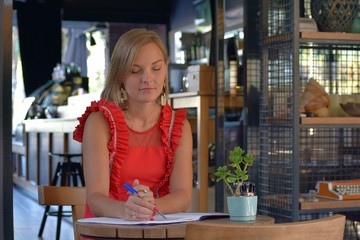A sitting blonde girl in a red dress in a cafe writes.