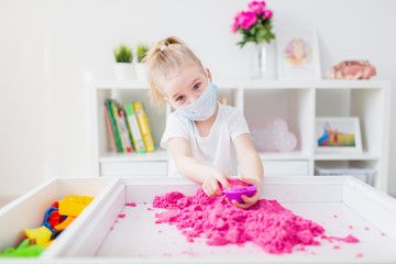 Home learning during virus pandemic. Little girl playing with pink kinetic sand at home, wearing...