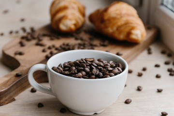 Cup full of coffee beans, two croissants on wooden board in the background