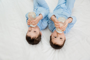 Two baby twin boys in pajamas lie on the bed drinking milk from bottles. View from above. concept...
