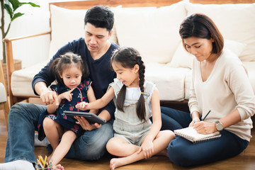 Young Asian love family stay in living room at home, father teaching two daughter to playing and use the  tablet by looking which sit on wooden floor near sofa, looking smiling and felling happy.