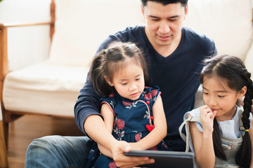 Young Asian love family stay in living room at home, father teaching two daughter to playing and use the  tablet by looking which sit on wooden floor near sofa, looking smiling and felling happy.