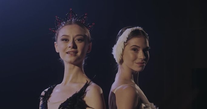 Portrait of Caucasian beautiful young female ballerinas in white and black dresses standing back to back and smiling to camera on dark background. Women in swans parts of ballet.