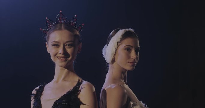Portrait of Caucasian beautiful young female ballerinas in white and black dresses and crown standing back to back and smiling to camera or looking at side. Women in swans parts of ballet.