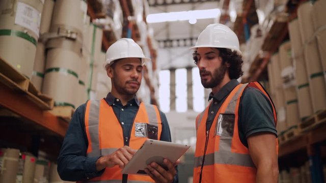Two male factory worker wearing white hardhat and uniform inspecting supplies in factory warehouse using digital tablet