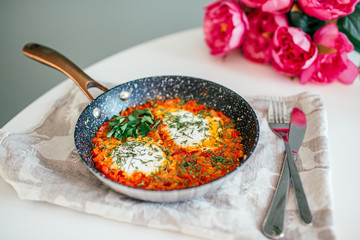 Shakshuka with eggs, bell pepper, tomato, and parsley in a cast iron pan on a white table with pink flowers on a background
