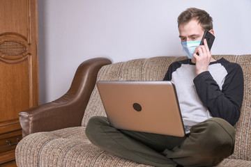 Young man in medical mask working at home in the room on the sofa using a laptop. Quarantine, self-isolation, coronavirus protection. Vacation from work.