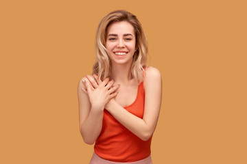 cheerful woman presses hands to chest and thanks, happy with generous gift, snow-white smile. Young woman with shoulder length blonde hair wears t-shirt isolated on orange background in Studio