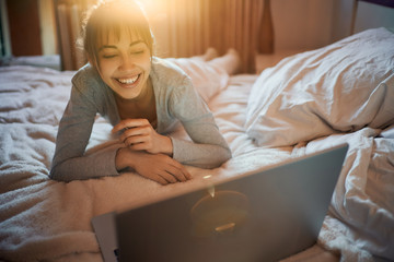 Cheerful girl having video chat with friends using laptop camera while lying on bed at home. Positive woman chatting in social networks on net-book device using wireless internet.