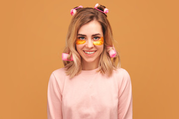 Pretty girl with patches on eyes and curlers on hair look at camera, has gentle smile, shows her natural beauty. Young woman with shoulder blonde wears pajamas isolated on orange background in Studio