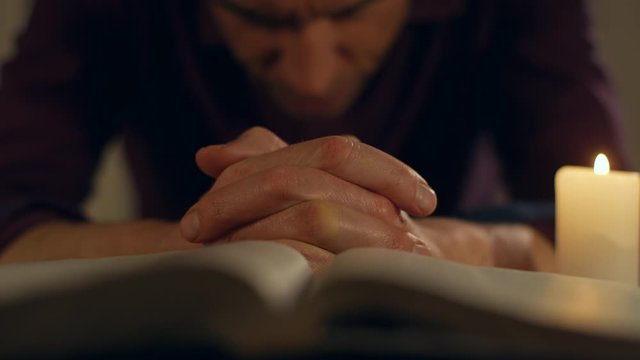 A man is sitting praying fervently at night with hands together in front of an open Bible and a candle light