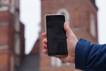 Hand of tourist holding mobile smartphone with black screen on the background of an old house, photographing sights or architecture. Journeys or Travel concepts and technology
