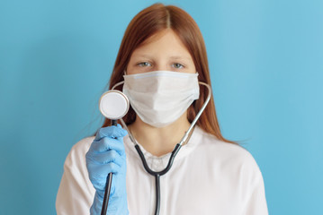 A teen girl plays doctor, a concept of choosing a profession for a child, a red hair girl in a white shirt, wearing a protective face mask and with a phonendoscope in her hands