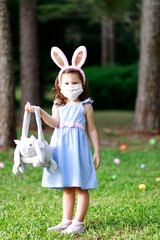 Little toddler girl with bunny ears and surgical face mask hunting for Easter eggs during coronavirus quarantine