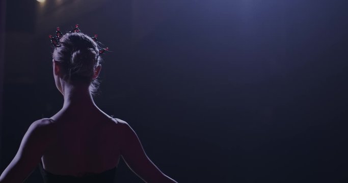 Back view on Caucasian slim graceful professional ballerina in black dress coming out on stage from darkness. Rear of female ballet dancer walking out the curtains and entering the stage to bow.