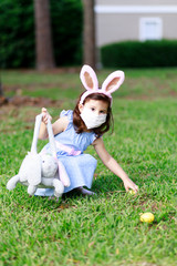 Little toddler girl with bunny ears and surgical face mask hunting for Easter eggs during coronavirus quarantine - 337056283