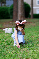 Little toddler girl with bunny ears and surgical face mask hunting for Easter eggs during coronavirus quarantine - 337056471