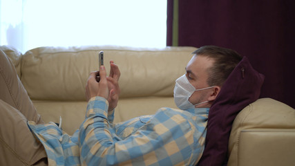 Obraz premium Man in a Medical Mask Uses a Smartphone at Home During Quarantine. Works Alone From Home Remotely. Quarantine Covid-19 Corona Virus. Social Distancing Concept.