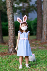 Little toddler girl with bunny ears and surgical face mask hunting for Easter eggs during coronavirus quarantine - 337056644